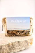 A beige fabric pouch with a printed front label reading 'SPA RITUAL SET' and 'MARIA', placed on a rough stone against a clean white background.