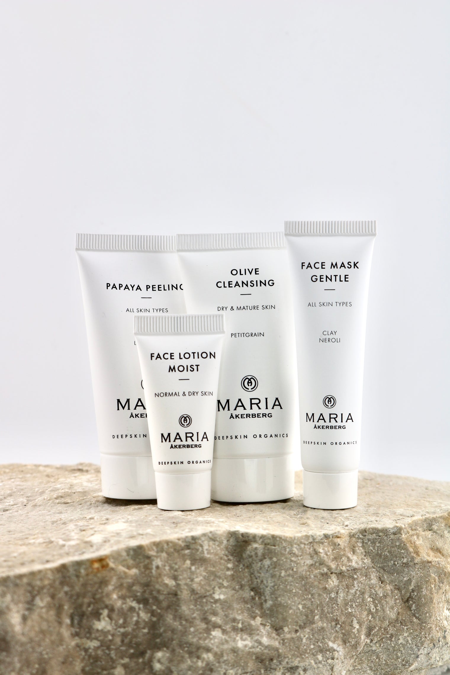 Four white skincare tubes from Maria Akenberg, including Face Lotion Moist, Olive Cleansing, Face Mask Gentle, and Papaya Peeling, arranged on a textured stone surface against a light gray background.